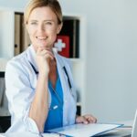 smiling female doctor with stethoscope looking away at table with clipboard and laptop in office
